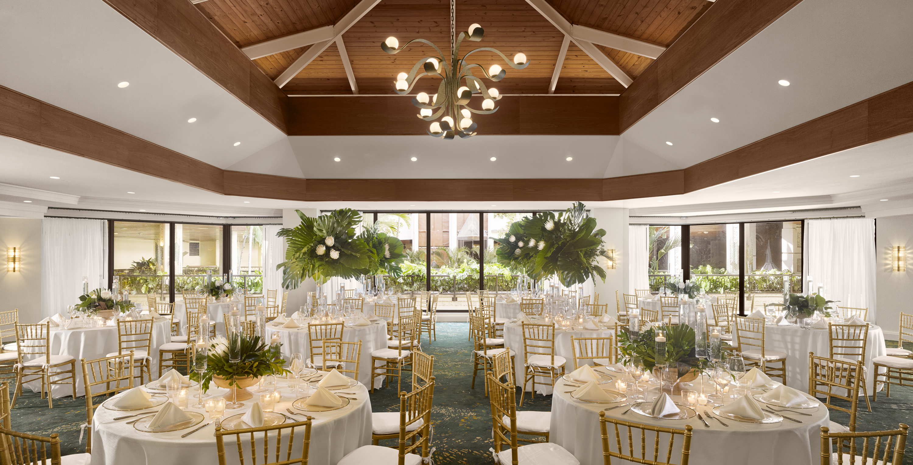 A large, elegantly set dining room with round tables, white tablecloths, gold chairs, and greenery centerpieces.