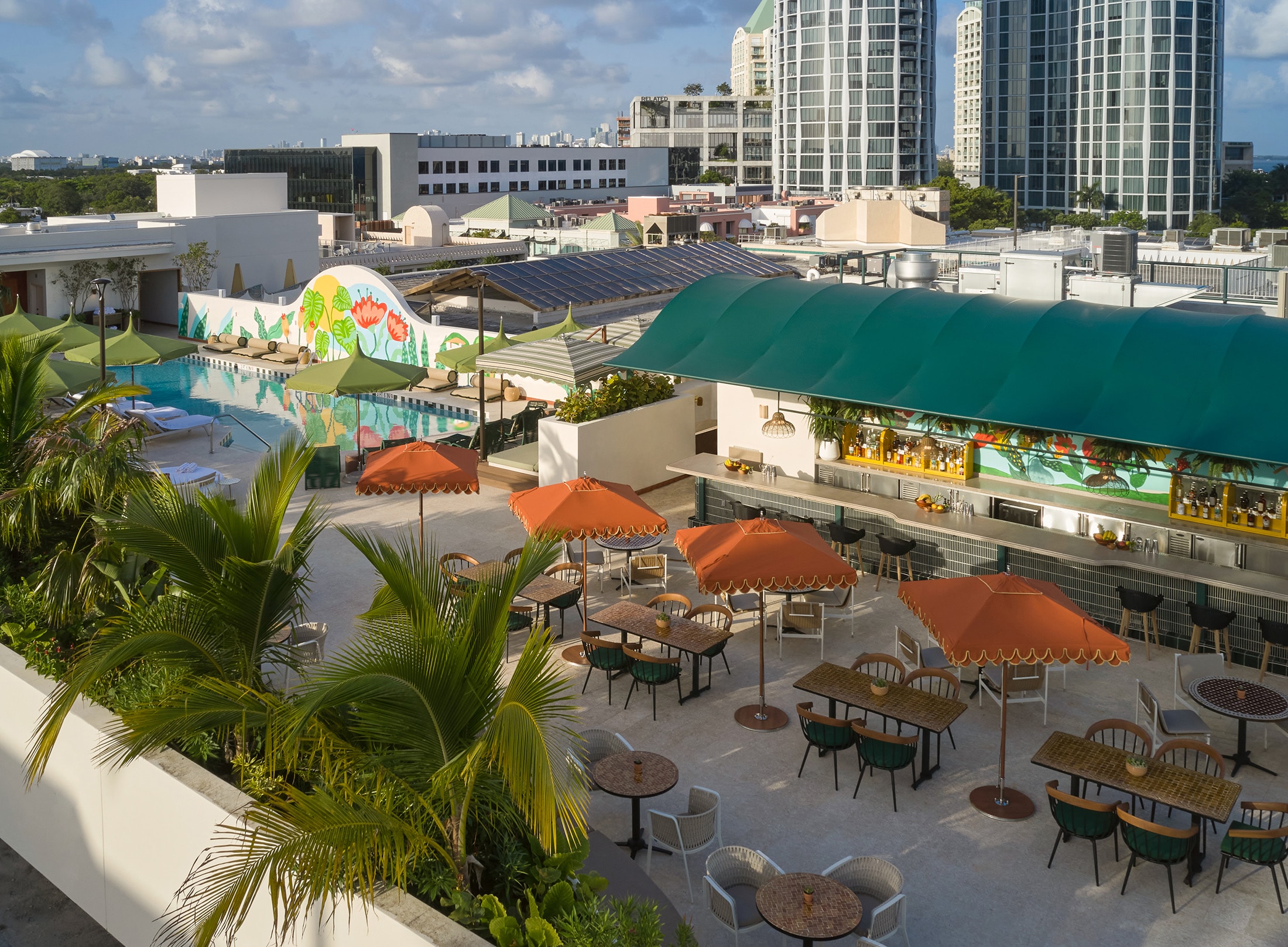 A rooftop pool and bar area with tables, umbrellas, and palm trees overlooking the city.