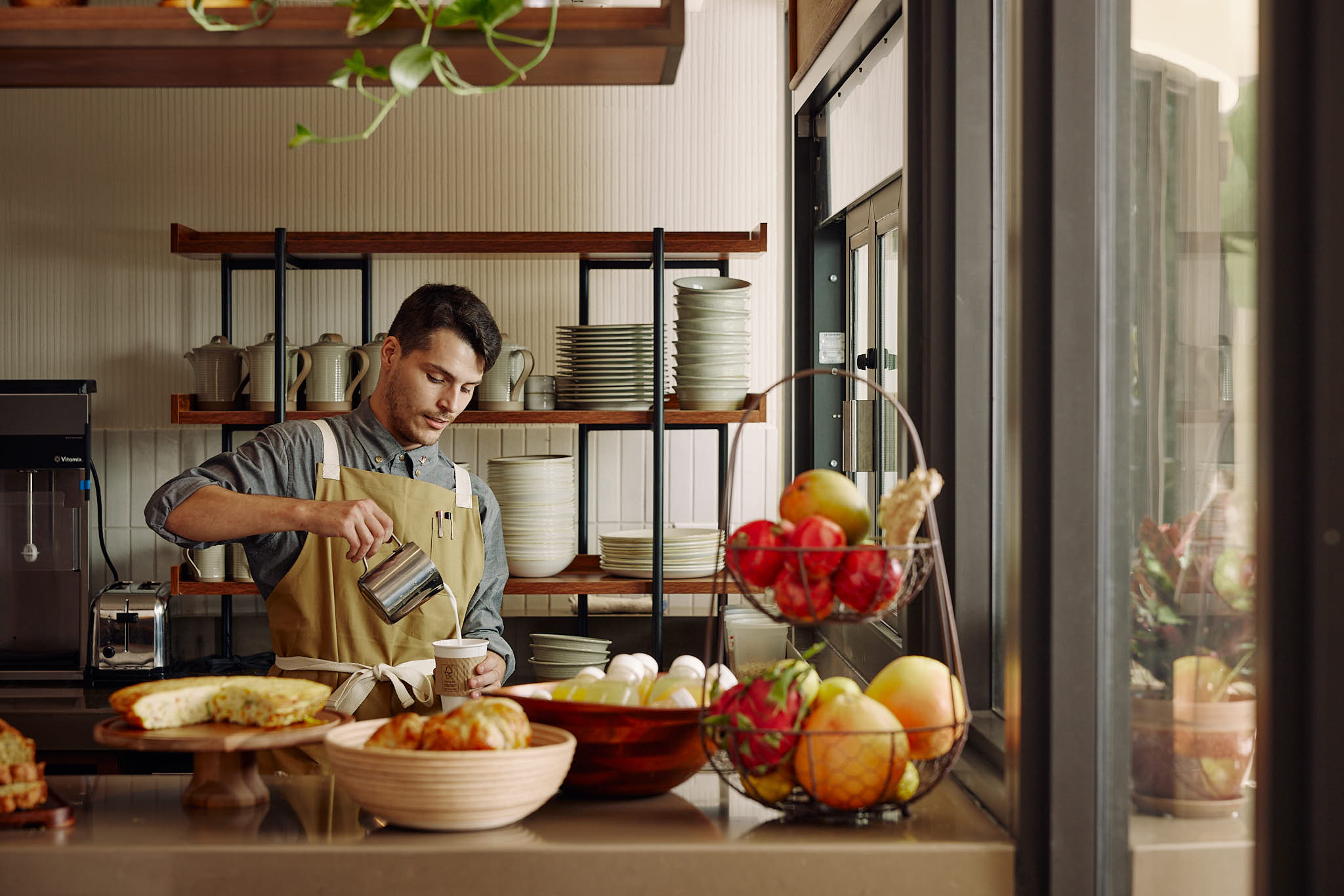 A man in an apron preparing a drink at a counter with pastries and fruit.