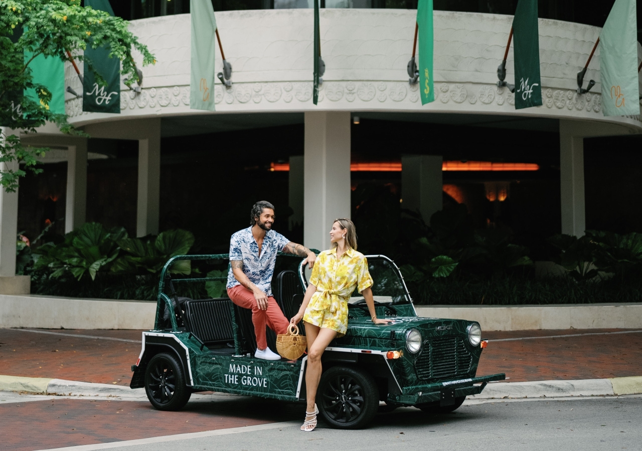 A couple poses with a green car in front of a white building with flags.