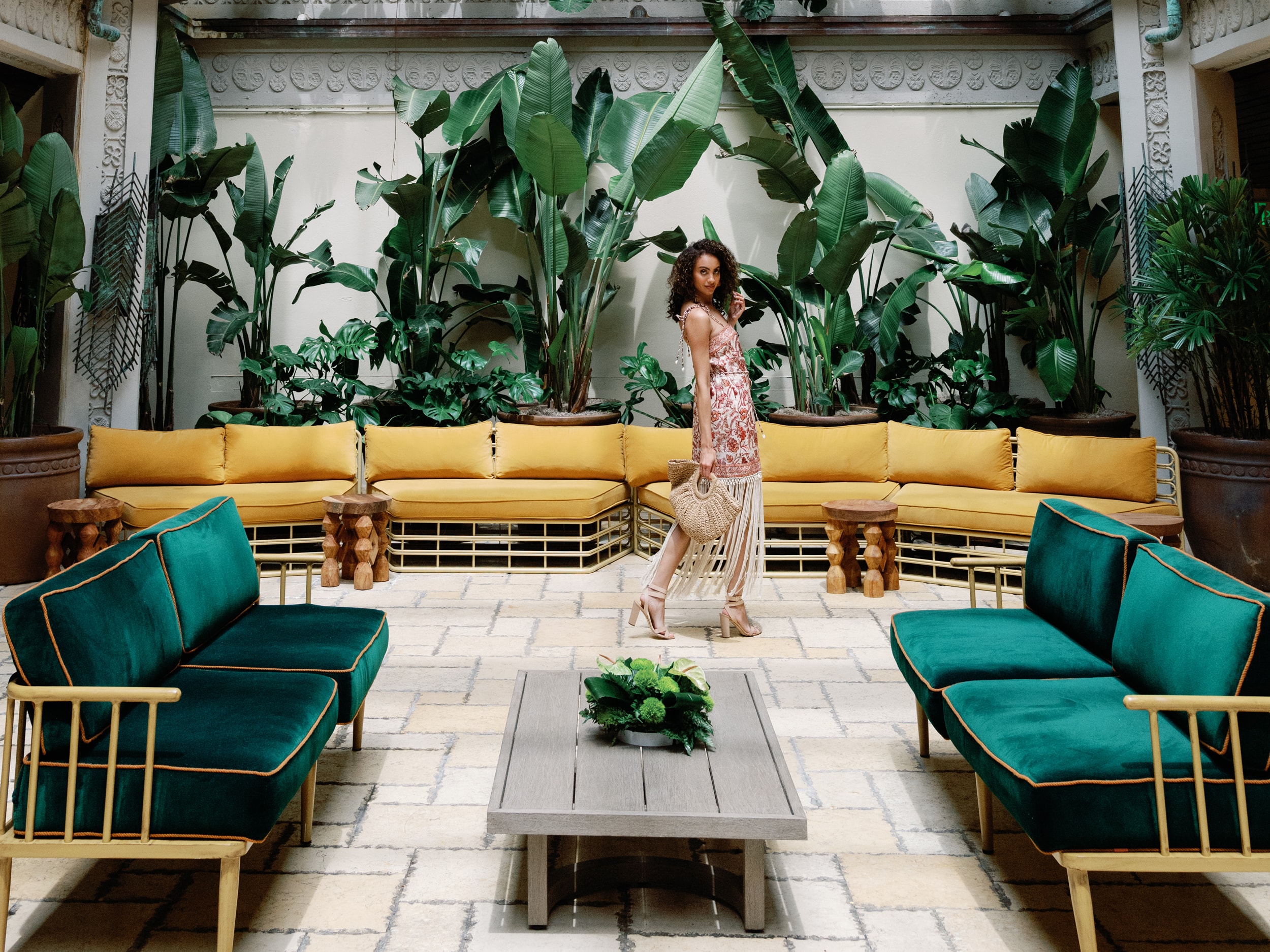 A woman standing in a lobby lounge with green sofas and yellow seating area surrounded by large plants.